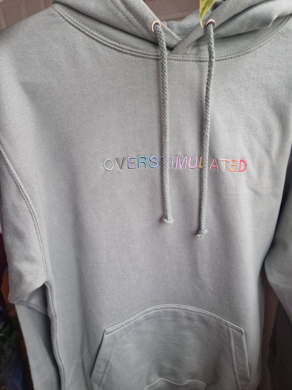 A light dusty green hoodie with the word 'OVERSTIMULATED' embroidered across the chest in pastel rainbow colours. The hoodie features matching dusty green drawstrings and a front kangaroo pocket, shown hanging against an indoor background.